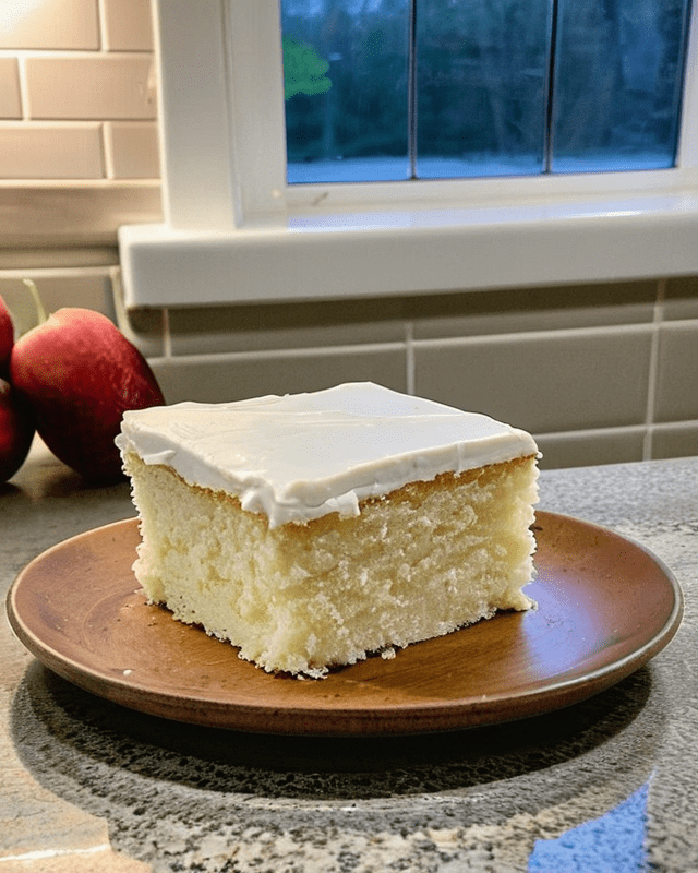 A slice of vanilla cake with vanilla frosting on a wooden plate in a kitchen.